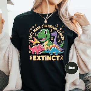 May include: Black t-shirt featuring a dinosaur graphic with the text "Let's Make Childhood Cancer Extinct". The design includes a green T-Rex, purple Brontosaurus, and other dinosaurs with yellow ribbons.