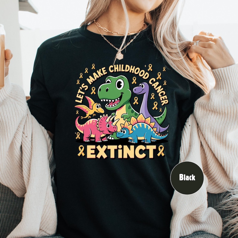 May include: Black t-shirt featuring a dinosaur graphic with the text "Let's Make Childhood Cancer Extinct". The design includes a green T-Rex, purple Brontosaurus, and other dinosaurs with yellow ribbons.