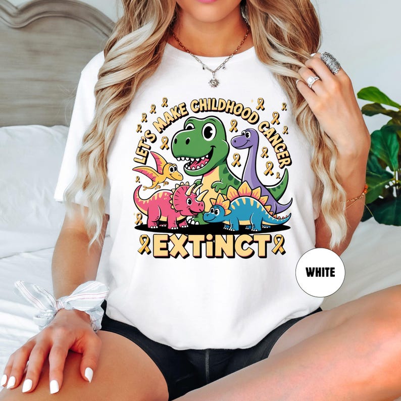 May include: White t-shirt with a dinosaur graphic and the text "Let's Make Childhood Cancer Extinct". The design features a green T-Rex, purple Brontosaurus, and other dinosaurs with yellow ribbons.