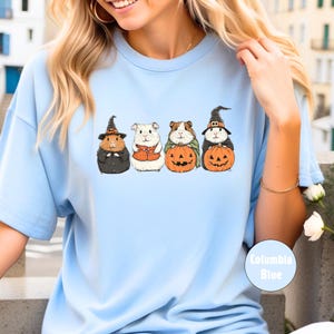 Pode incluir: Camiseta azul claro com um gr&aacute;fico de quatro porquinhos-da-&iacute;ndia vestidos com fantasias de Halloween. Os porquinhos-da-&iacute;ndia est&atilde;o usando chap&eacute;us de bruxa, um colete e segurando ab&oacute;boras.