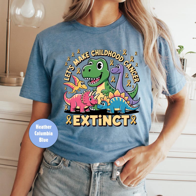 May include: Heather Columbia blue t-shirt with a colourful dinosaur graphic. The design includes the text "Let's Make Childhood Cancer Extinct" with yellow ribbons. The shirt has a crew neck and short sleeves.