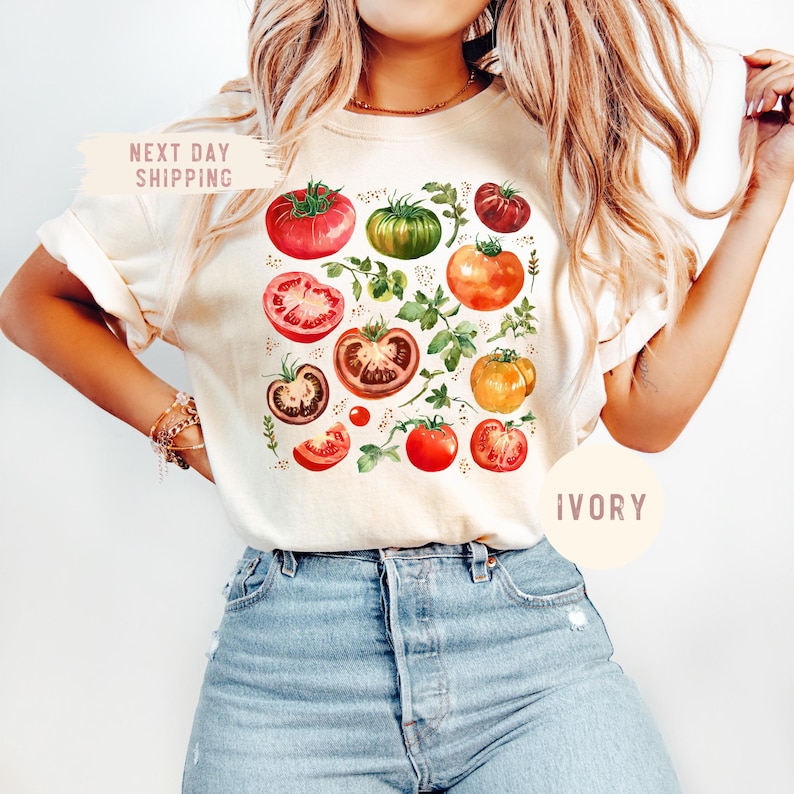 May include: Ivory-colored t-shirt featuring a watercolor illustration of various tomatoes and green foliage. The design includes whole and sliced tomatoes in shades of red, orange, and green. The text "NEXT DAY SHIPPING" is visible on the top left.