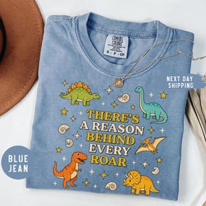 May include: Blue jean Comfort Colors t-shirt with a dinosaur graphic and the text "THERE'S A REASON BEHIND EVERY ROAR." The shirt features illustrations of various dinosaurs in orange, green, and yellow. A gold necklace is draped across the shirt.