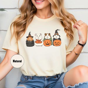 Pode incluir: Uma camiseta amarela clara com um gr&aacute;fico de quatro porquinhos-da-&iacute;ndia vestidos com fantasias de Halloween. Os porquinhos-da-&iacute;ndia est&atilde;o segurando ab&oacute;boras e usando chap&eacute;us de bruxa. O texto "Natural" est&aacute; impresso no canto inferior esquerdo da camiseta.