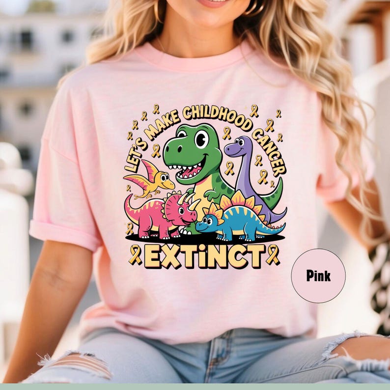 May include: Pale pink t-shirt with a dinosaur graphic and the words "Let's Make Childhood Cancer Extinct". The design features a green T-Rex, purple Brontosaurus, and other dinosaurs with yellow ribbons.