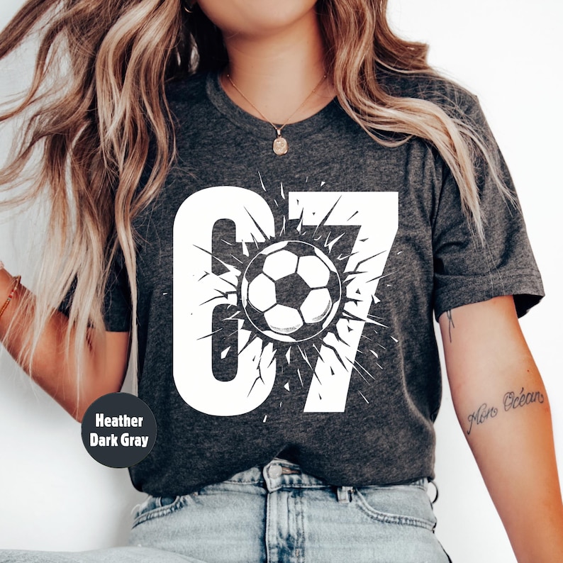 May include: Heather dark gray t-shirt with a large white graphic of the number 67 and a soccer ball. The soccer ball is in the center of the number 67 and has a burst design around it.