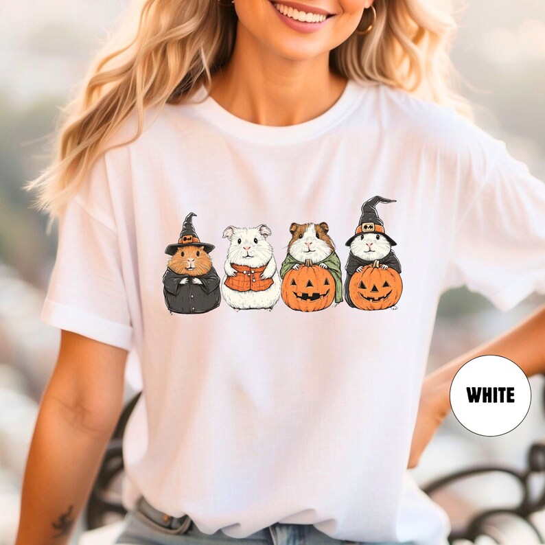 Pode incluir: Camiseta branca com um gr&aacute;fico de quatro porquinhos-da-&iacute;ndia vestidos com fantasias de Halloween segurando ab&oacute;boras. Os porquinhos-da-&iacute;ndia est&atilde;o usando chap&eacute;us de bruxa, uma camisa xadrez e um chap&eacute;u de bruxa. O texto "WHITE" &eacute; vis&iacute;vel no canto inferior direito da imagem.