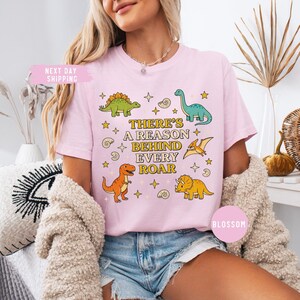 May include: Light pink t-shirt featuring colorful dinosaur illustrations and the text "THERE'S A REASON BEHIND EVERY ROAR." The design includes a green Stegosaurus, a blue Brontosaurus, and an orange T-Rex. The shirt has a relaxed fit.
