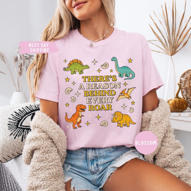 May include: Light pink t-shirt featuring colorful dinosaur illustrations and the text "THERE'S A REASON BEHIND EVERY ROAR." The design includes a green Stegosaurus, a blue Brontosaurus, and an orange T-Rex. The shirt has a relaxed fit.