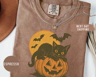 Pode incluir: Camiseta cor espresso com design de Halloween. O gráfico mostra um gato preto em uma abóbora laranja, com uma lua amarela e morcegos. A camiseta tem gola redonda e mangas curtas.