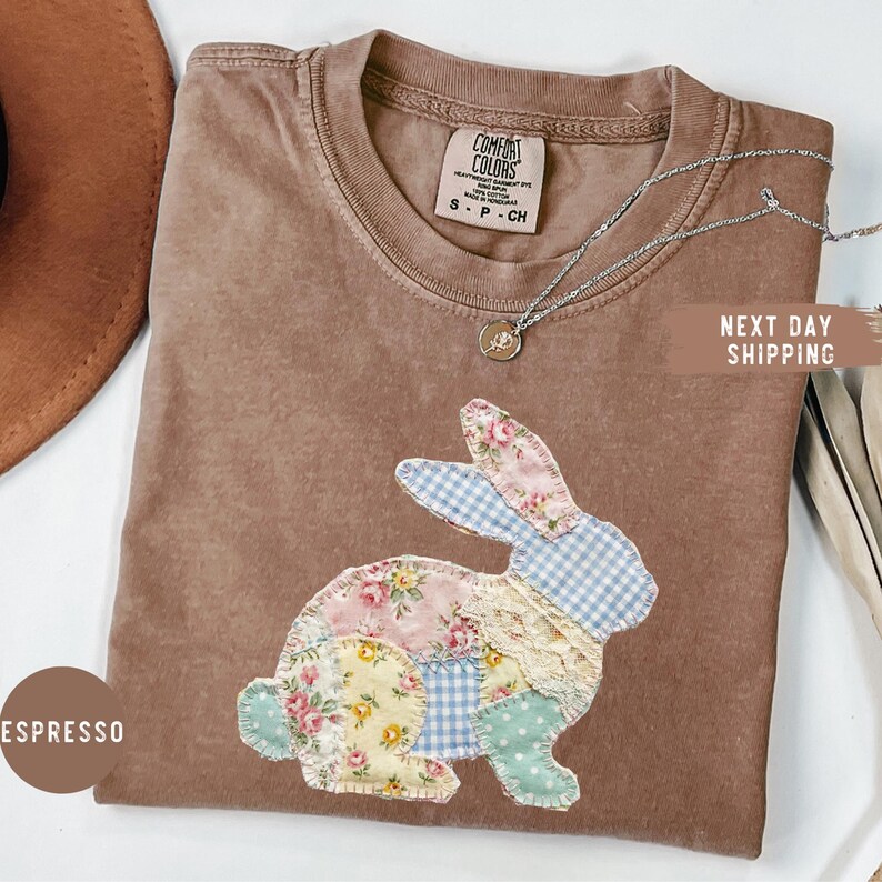 May include: Espresso-colored Comfort Colors t-shirt with a patchwork bunny design. The bunny is made of floral, gingham, and lace fabrics. A silver necklace with a small pendant is on the shirt. The shirt is labeled S-P-CH.
