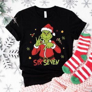 K&ouml;nnte beinhalten: Schwarzes T-Shirt mit einem gr&uuml;nen Grinch-Charakter in einem Weihnachtsmann-Anzug, der eine Handbewegung macht. Der Text "SIX SEVEN" ist in Gr&uuml;n und Gelb gedruckt. Festliches Design mit Schneeflocken und Sternen.