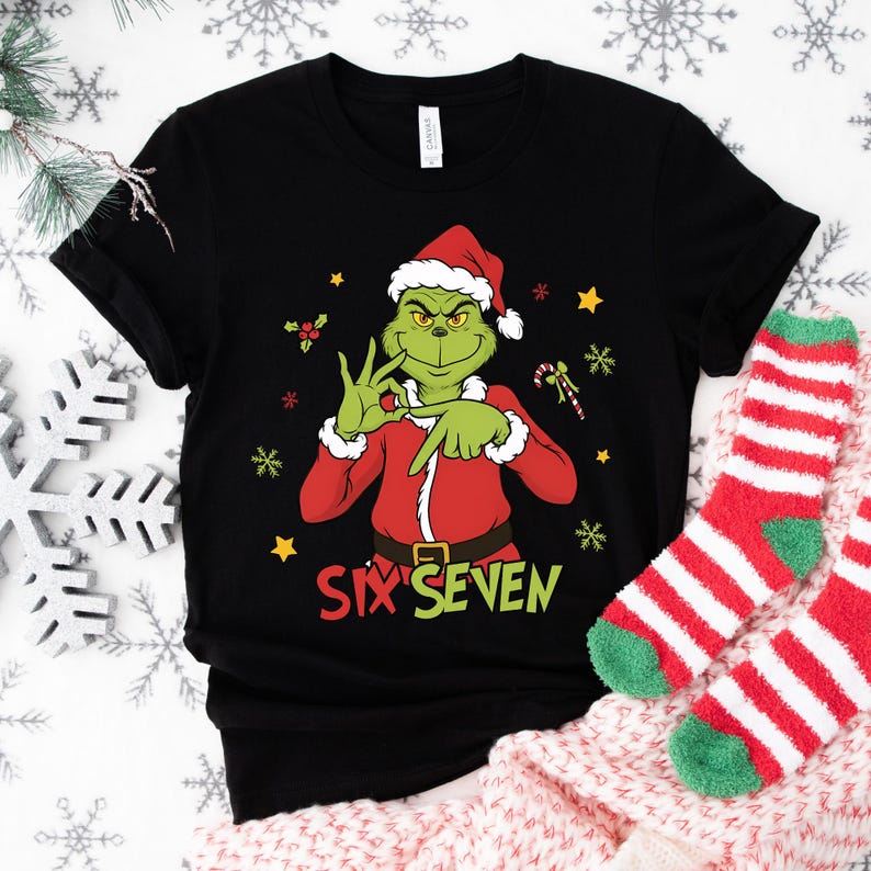 K&ouml;nnte beinhalten: Schwarzes T-Shirt mit einem gr&uuml;nen Grinch-Charakter in einem Weihnachtsmann-Anzug, der eine Handbewegung macht. Der Text "SIX SEVEN" ist in Gr&uuml;n und Gelb gedruckt. Festliches Design mit Schneeflocken und Sternen.