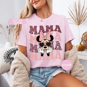 May include: Pink t-shirt with the word "MAMA" in a leopard print design. The shirt features a Minnie Mouse graphic wearing heart-shaped sunglasses and a bow. The shirt is a light pink color.