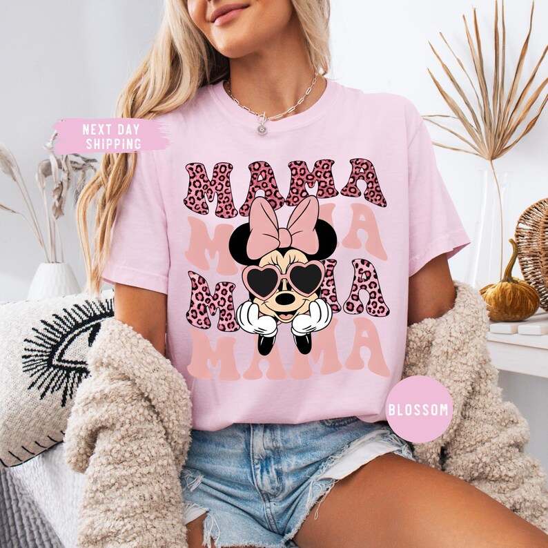 May include: Pink t-shirt with the word "MAMA" in a leopard print design. The shirt features a Minnie Mouse graphic wearing heart-shaped sunglasses and a bow. The shirt is a light pink color.