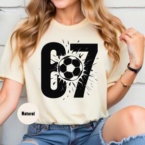 May include: A light yellow t-shirt featuring a large black graphic of the number 67 with a soccer ball in the center. The soccer ball is black and white. The word "Natural" is printed on the bottom left of the shirt.