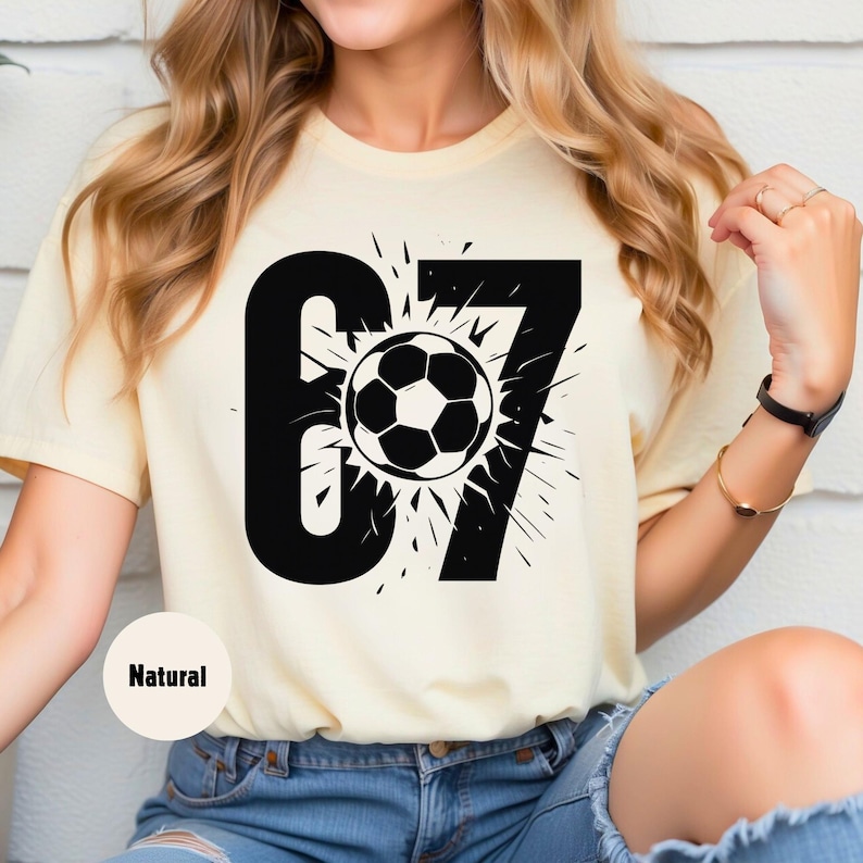 May include: A light yellow t-shirt featuring a large black graphic of the number 67 with a soccer ball in the center. The soccer ball is black and white. The word "Natural" is printed on the bottom left of the shirt.