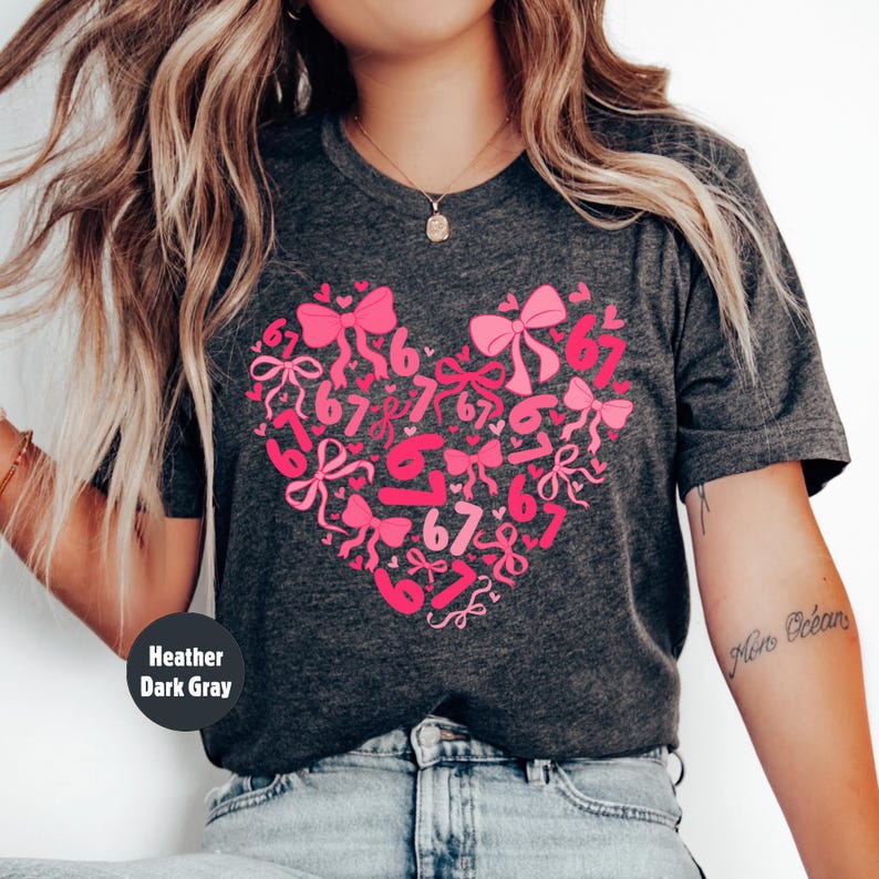 May include: Heather dark gray t-shirt with a pink heart design. The heart is filled with pink bows and the number "67". The shirt is a crew neck and has short sleeves. The text "Heather Dark Gray" is in a circle on the bottom left.