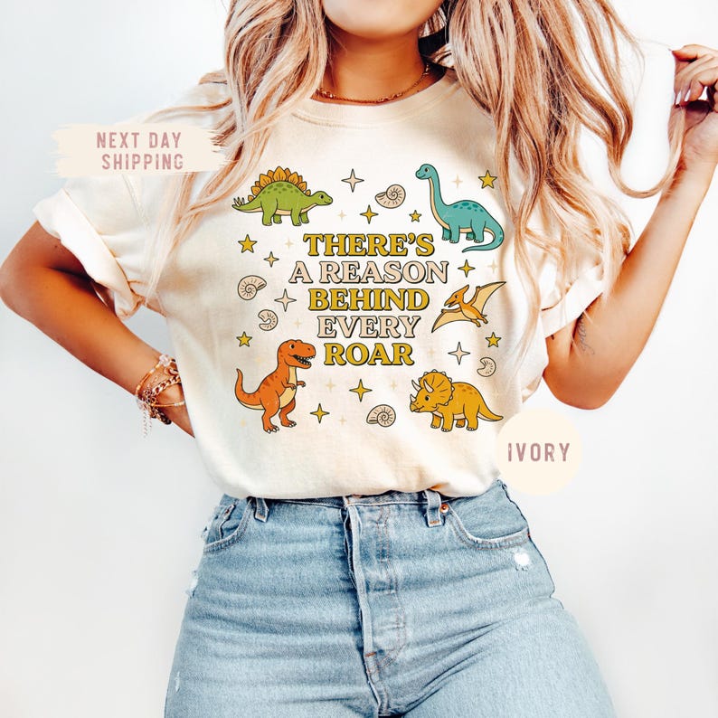 May include: Ivory-colored t-shirt featuring colorful dinosaur illustrations and the text "THERE'S A REASON BEHIND EVERY ROAR." The design includes a green Stegosaurus, a blue Brontosaurus, an orange T-Rex, and a yellow Triceratops, along with stars and shell graphics.