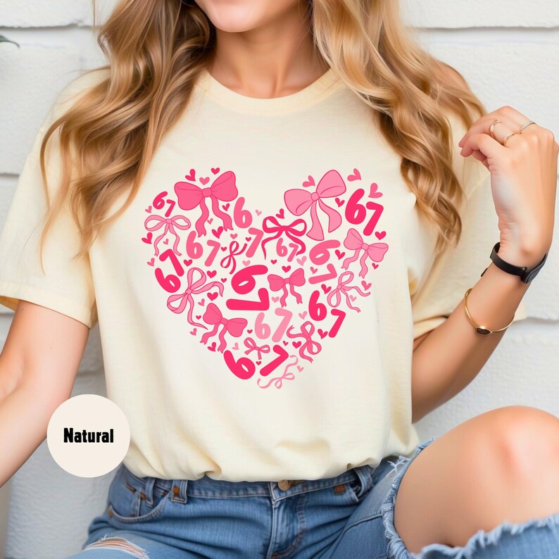 May include: A light yellow t-shirt featuring a heart-shaped design filled with pink bows and the number "67". The shirt has a relaxed fit and is made of a soft material. The word "Natural" is printed on the shirt.