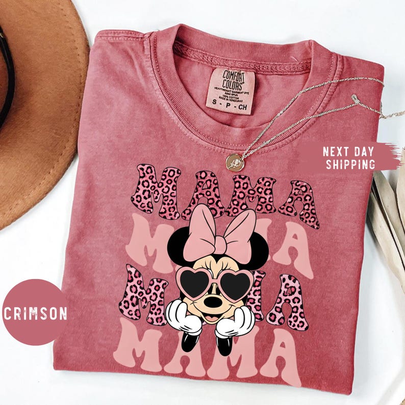 May include: Crimson-colored t-shirt with the word "MAMA" in a pink, leopard print design. The shirt features a cartoon character with heart-shaped sunglasses and a pink bow. The shirt is a Comfort Colors brand.