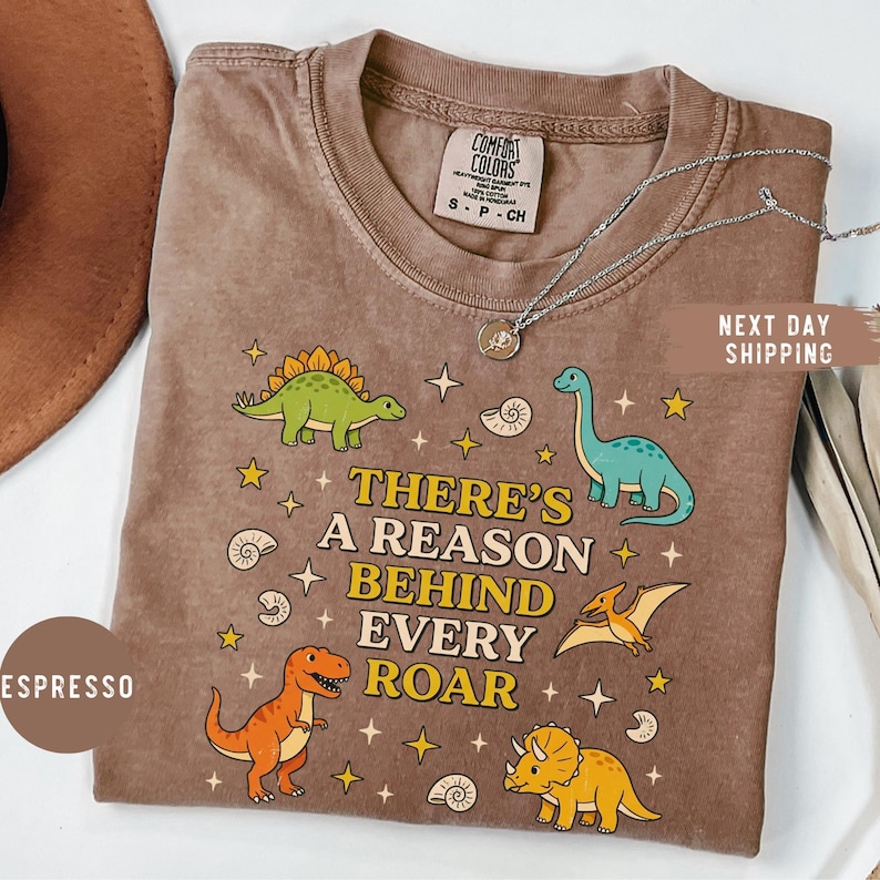 May include: Espresso-colored t-shirt with a dinosaur graphic and the text "THERE'S A REASON BEHIND EVERY ROAR." The shirt features illustrations of various dinosaurs, stars, and shells. The shirt is a Comfort Colors brand.