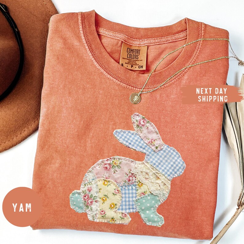 May include: A coral-colored t-shirt featuring a patchwork bunny design. The bunny is made of floral, gingham, and lace fabrics. A gold necklace with a pendant rests on the shirt. The shirt has a Comfort Colors tag.