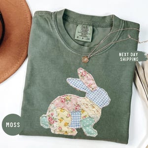May include: Moss green t-shirt with a patchwork bunny design. The bunny is made of floral, gingham, and lace fabrics. A gold necklace with a pendant is draped on the shirt. The text "NEXT DAY SHIPPING" is visible.