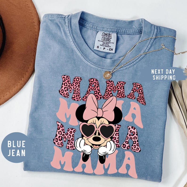 May include: Blue jean Comfort Colors t-shirt with the word "MAMA" in pink leopard print and a Minnie Mouse graphic wearing heart-shaped sunglasses. A gold necklace is draped across the shirt.
