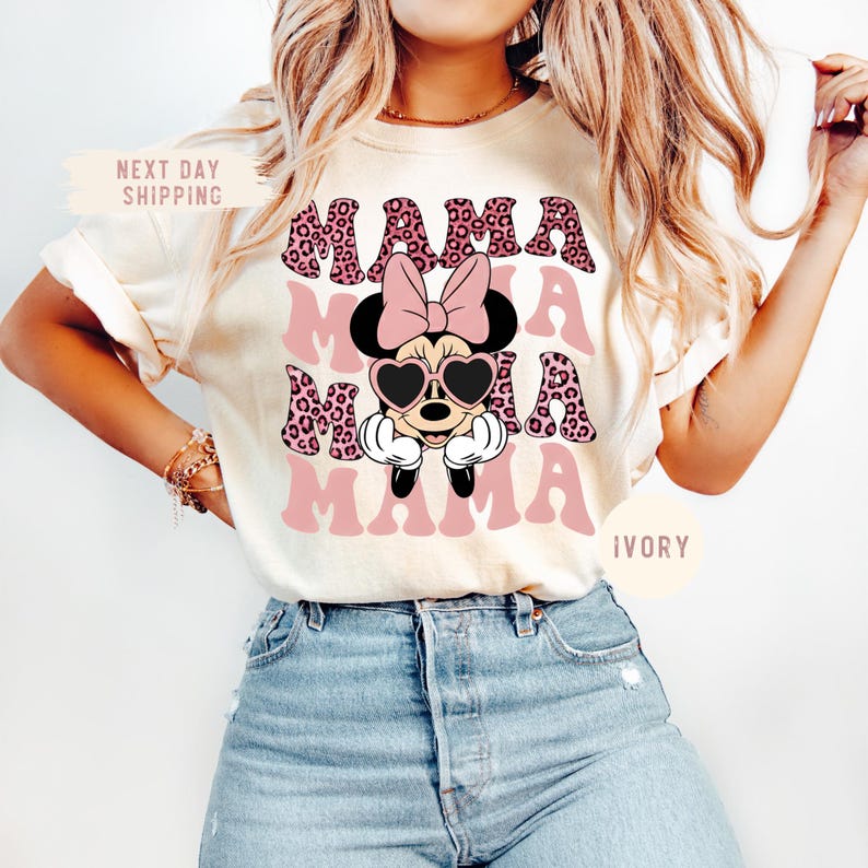 May include: Ivory-colored t-shirt featuring the word "MAMA" in a pink leopard print design, repeated three times. A Minnie Mouse graphic with heart-shaped sunglasses and a pink bow is centered on the shirt. The shirt has short sleeves.