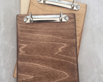 May include: Three wooden clipboards with silver binder clips. The clipboards are stacked on top of each other. The top clipboard is light brown, the middle clipboard is medium brown, and the bottom clipboard is dark brown.