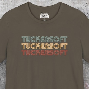 Könnte beinhalten: Olivgrünes T-Shirt mit dem Wort "TUCKERSOFT" dreifach in Türkis, Gold und Rot übereinander angeordnet. Das Shirt hat einen Rundhalsausschnitt und kurze Ärmel. Das Markenlogo ist oben sichtbar.