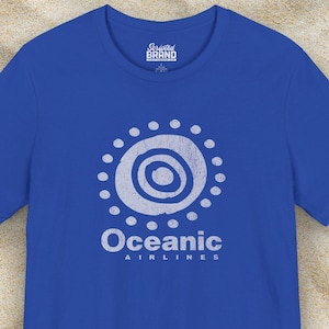 May include: A royal blue t-shirt featuring a white logo. The logo is a circle with a smaller circle inside, surrounded by dots. The text "Oceanic Airlines" is below the logo.