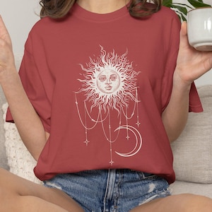 Op de afbeelding: Een baksteenrood T-shirt met korte mouwen en een witte grafische print van een zon met een menselijk gezicht, hangende kettingen en een maansikkel. Het T-shirt heeft een ronde hals en een relaxte pasvorm met een centraal ontwerp.