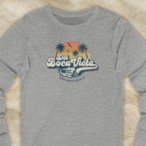 May include: A heather grey long-sleeve t-shirt with a vintage-style graphic of a palm tree sunset and the text "Del Boca Vista" with a retro camper van underneath. The text below the graphic reads "Where people come to die."