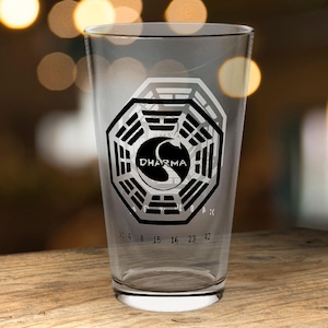 May include: A clear pint glass featuring the Dharma Initiative logo in black and silver, with the numbers 4 8 15 16 23 42. The glass is displayed on a wooden surface.
