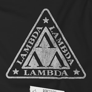 Op de afbeelding: Zwart t-shirt met een verweerd wit driehoekontwerp. De driehoek heeft het woord "LAMBDA" langs de randen en twee Lambda-symbolen binnenin. Een klein label onderaan zegt "VINTAGE DISTRESSED TEES".