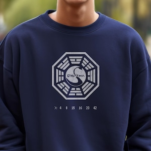 May include: A navy blue sweatshirt featuring a white Dharma Initiative logo and numbers. The logo is a circle with a white swan inside and the word "DHARMA" written in white.