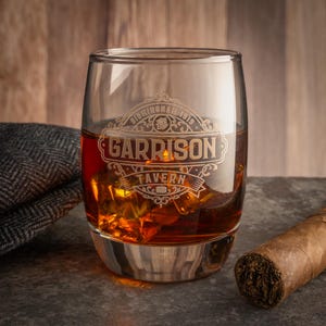Op de afbeelding: Een helder whiskyglas gevuld met amberkleurige vloeistof en ijs. Het glas is geëtst met de woorden "Garrison Tavern" en "Birmingham 1919". Een tweed pet en een sigaar liggen naast het glas.