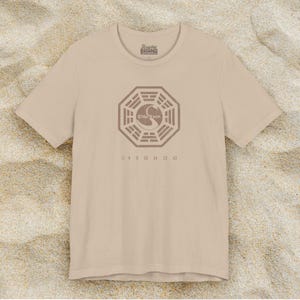 Tan short-sleeve t-shirt featuring the Dharma Initiative swan logo graphic from the TV show LOST in a vintage distressed style.