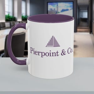 May include: A white ceramic mug with a purple interior and handle. The mug features the text "Pierpoint & Co" in a purple font, along with a geometric logo. The mug is sitting on a light gray surface.