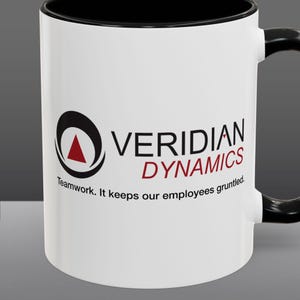 May include: White ceramic mug with a black rim and handle. The mug features a black and red logo with the text "VERIDIAN DYNAMICS" and the tagline "Teamwork. It keeps our employees gruntled."