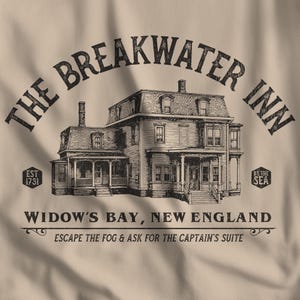 以下が含まれることがあります： ベージュ色のグラフィックTシャツ。The Breakwater Innの詳細なイラストが描かれており、「Widow's Bay, New England」の文字があります。デザインには「Escape the Fog & Ask for the Captain's Suite」と「By the Sea」のフレーズが含まれています。