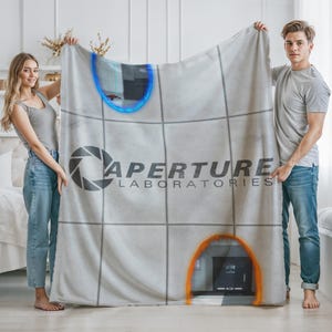 May include: A light gray blanket featuring the Aperture Laboratories logo and portal designs. The blanket has a grid pattern and the text "APERTURE LABORATORIES" in gray. Two people are holding the blanket.