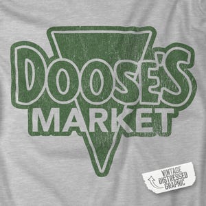 Könnte beinhalten: Graues T-Shirt mit einem verblichenen grünen Aufdruck. Der Aufdruck zeigt "DOOSE'S MARKET" in einer Vintage-Schriftart, eingebettet in ein grünes Dreieck. Ein kleines Etikett unten rechts zeigt "VINTAGE DISTRESSED GRAPHIC".