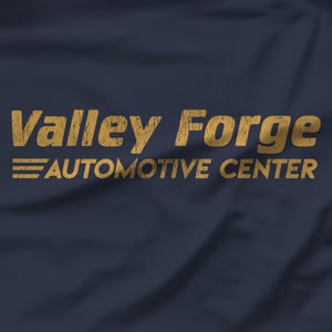 Puede incluir: Un fondo de tela azul marino oscuro con el texto de color dorado Valley Forge AUTOMOTIVE CENTER con una textura desgastada y envejecida. A la izquierda del texto hay tres líneas horizontales, también en un acabado dorado desgastado.