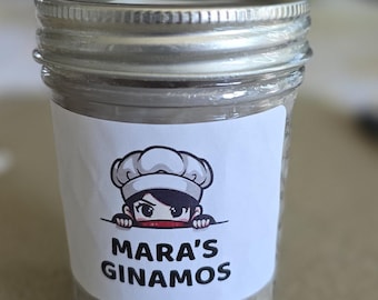 Könnte beinhalten: Ein klares Glas mit silbernem Deckel, gefüllt mit einer körnigen Substanz. Ein weißes Etikett auf dem Glas zeigt einen Cartoon-Koch und den Text "MARA'S GINAMOS". Das Glas steht auf einer hellbraunen Oberfläche.