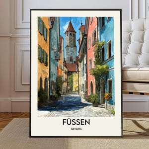 May include: A framed art print depicting a street scene in Fussen, Bavaria. The image shows vibrant buildings in orange, blue, and red along a cobbled street. A tall tower with a red roof is in the background. The text "FÜSSEN" and "BAVARIA" are at the bottom of the print.