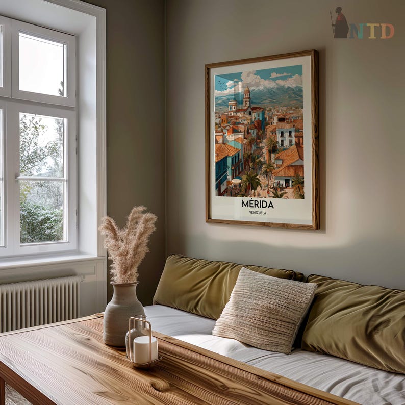 K&ouml;nnte beinhalten: Ein gerahmter Kunstdruck von M&eacute;rida, Venezuela, mit einer farbenfrohen Stadtlandschaft mit roten D&auml;chern, blauen Geb&auml;uden und einem Bergpanorama. Der Druck befindet sich in einem Holzrahmen und h&auml;ngt &uuml;ber einem Sofa mit olivgr&uuml;nen Kissen.