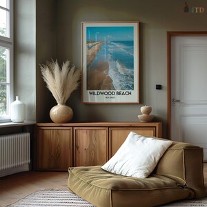 May include: A framed print of Wildwood Beach, New Jersey, with a blue sky and ocean. The print hangs above a wooden cabinet with a vase of pampas grass and a decorative bowl. A green floor cushion with a white pillow is in the foreground.